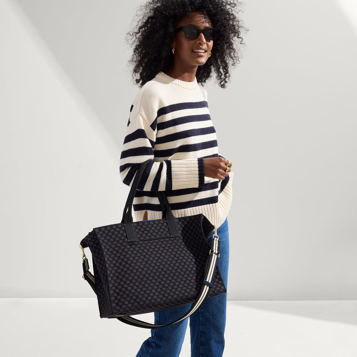 Overlay:: Shown with The Crossbody Strap (sold separately.) | Alt:: The Classic Tote in Black Sand, carried by its top handles by a model, shown from side.